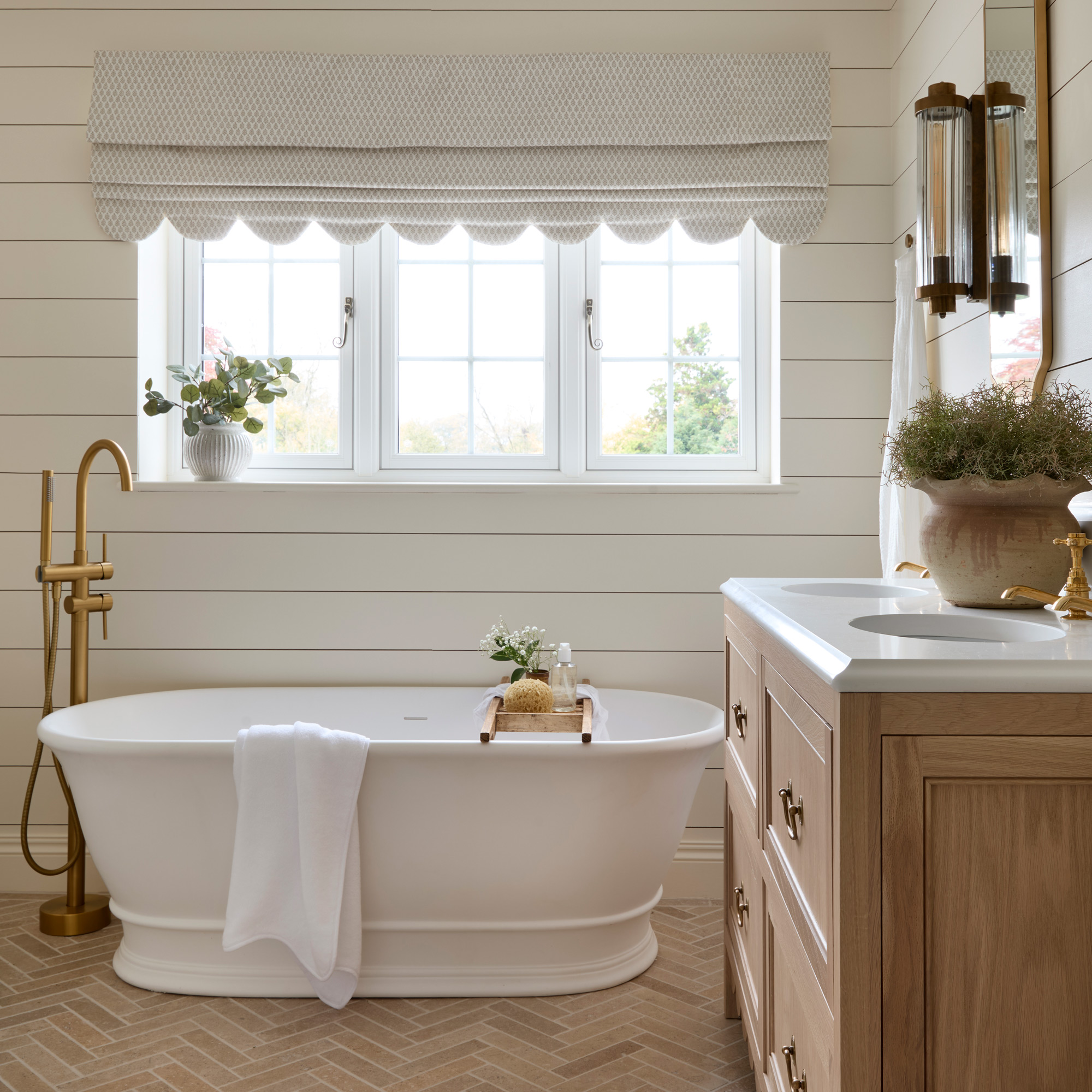 a neutral bathroom with horizontal wall panelling, a large white freestanding bath tub and brass fixtures, double vanity unit, a scalloped edge roman blind and limestone herringbone floor tiles