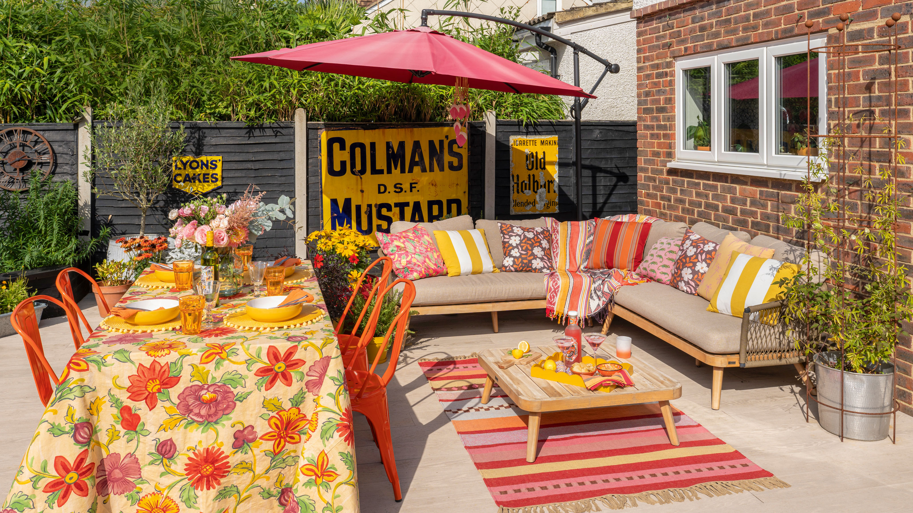 Patio with beige outdoor corner sofa, pink umbrella, orange garden chairs and floral table cloth