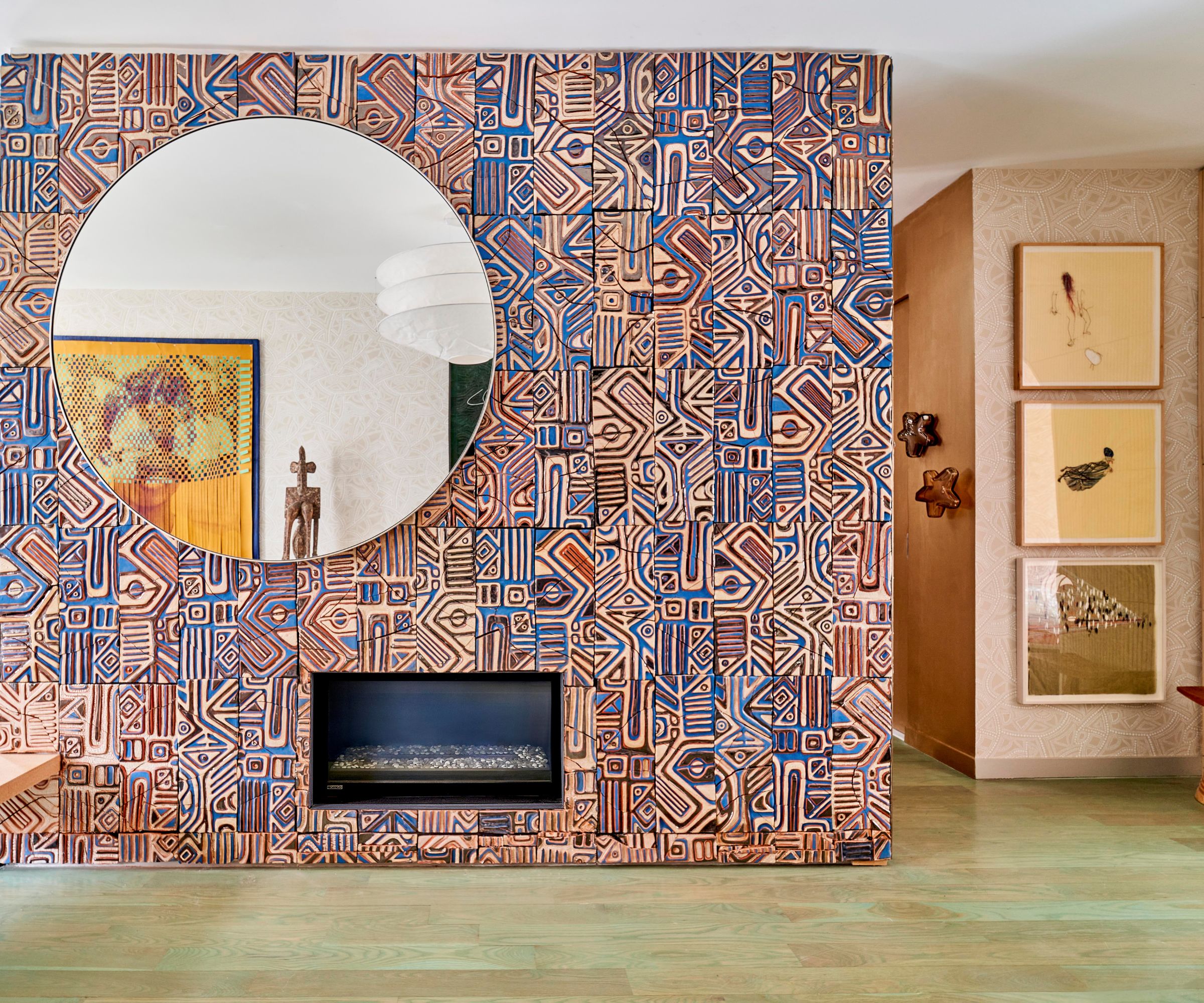 A modern fireplace wall covered in blue and brown patterned, geometric tiles, featuring a large circular mirror and light green wood flooring