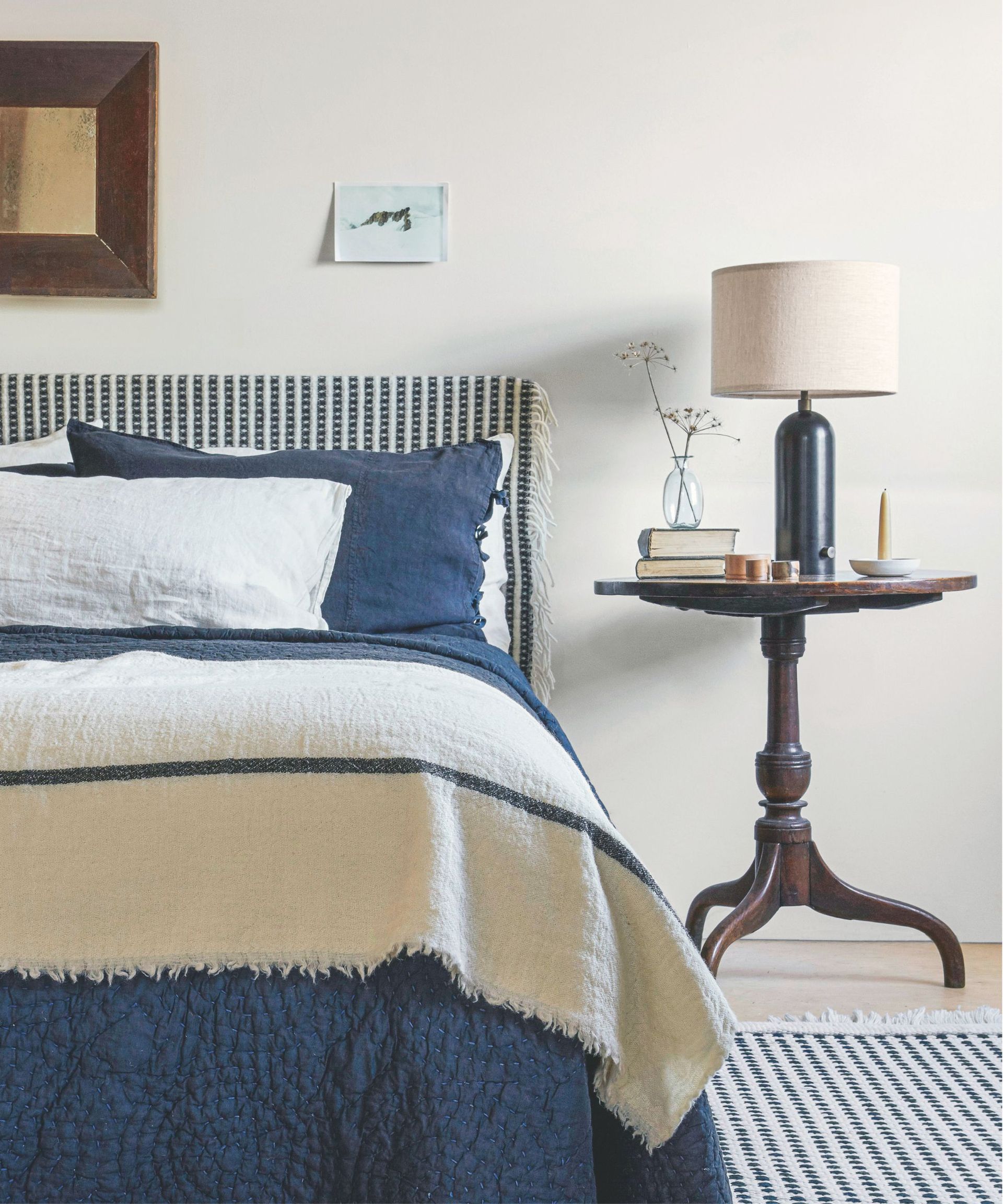 A blue and white bedroom with a blue and white bed with matching headboard and rug, a wooden bedside table with books, a vase with candles and a black and ecru lamp, and a framed gold picture in a wooden frame above the bed.