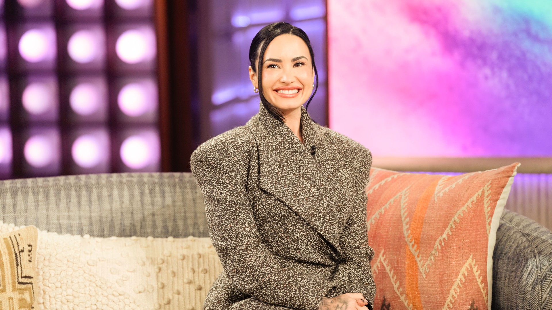 a young woman with dark hair sits on a couch on the set of a talk show