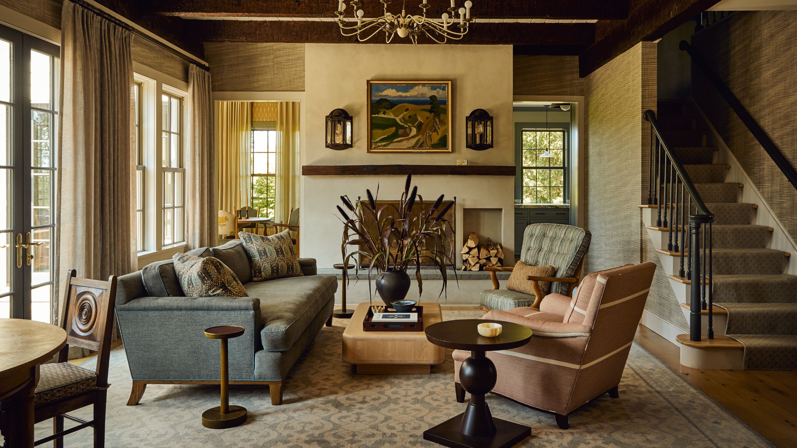 a cozy wood clad living room with exposed beams, plaster fireplace. french doors and windows, slate blue sofa and mismatched armchairs with the stairs in the corner