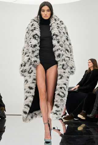 The leopard print coat trend is shown on the fall runway 2024 collection of Alaïa with a model walking wearing a white leopard coat over a black body suit with blue ankle strap pumps