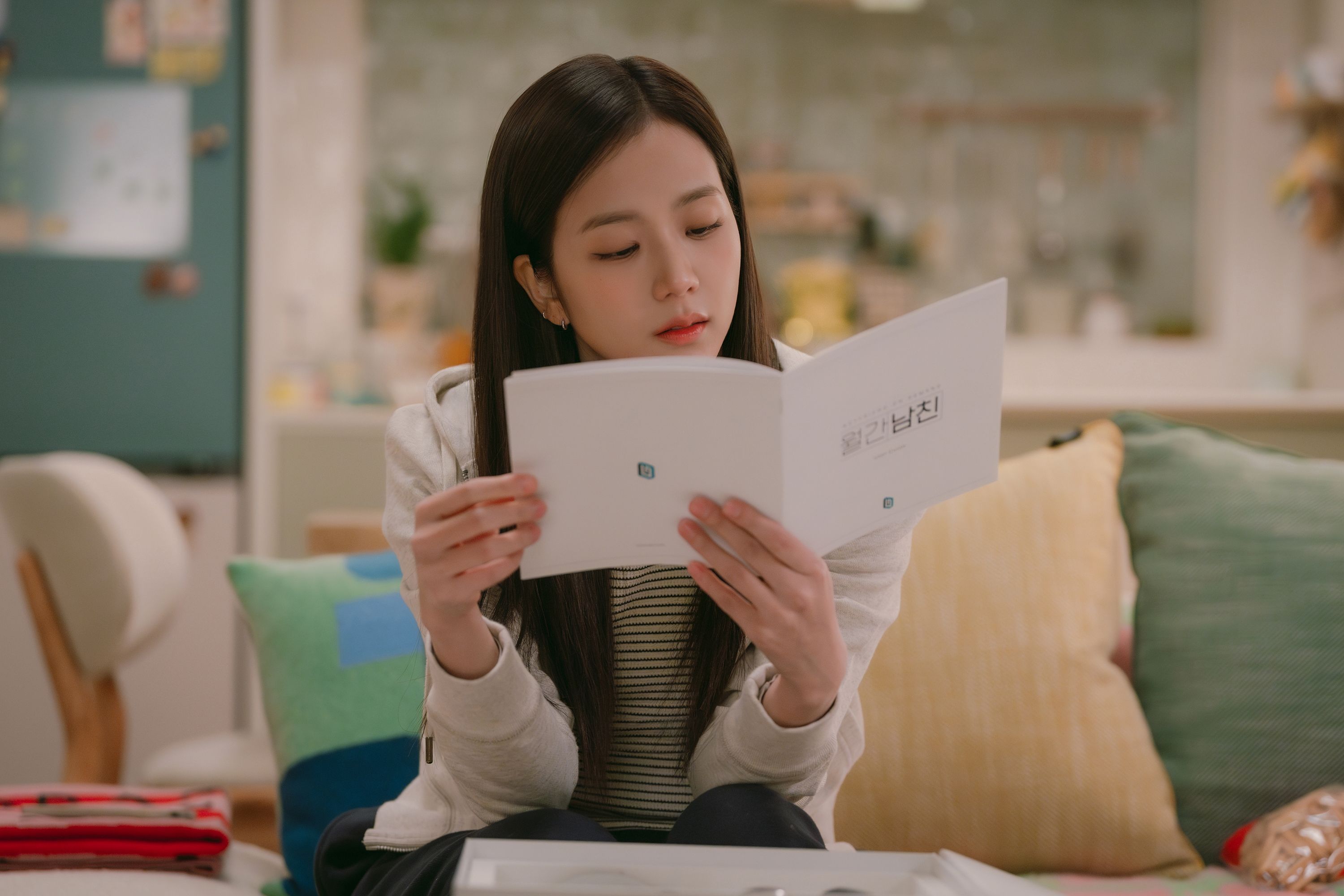 A woman (Jisoo of Blackpink) reads a Korean device manual while sitting on a couch in an apartment, in the Netflix K-drama 'Boyfriend on Demand.'