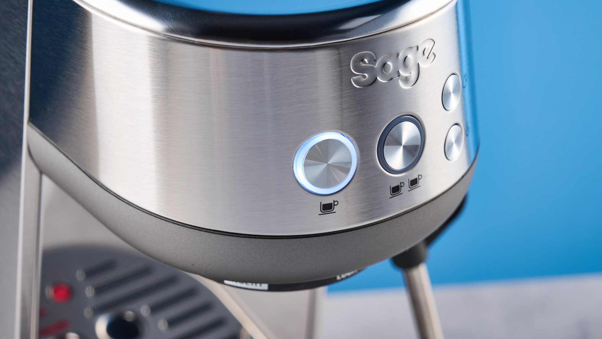 the breville bambino / sage bambino in silver photographed against a blue background with a wooden feature wall, showing the steam wand, group head, water tank, button controls, and espresso