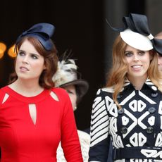 Princess Eugenie wears a bright red dress and a black ribbon on her head, while her sister Princess Beatrice wears a black and white patterned dress with a white hat featuring a black bow