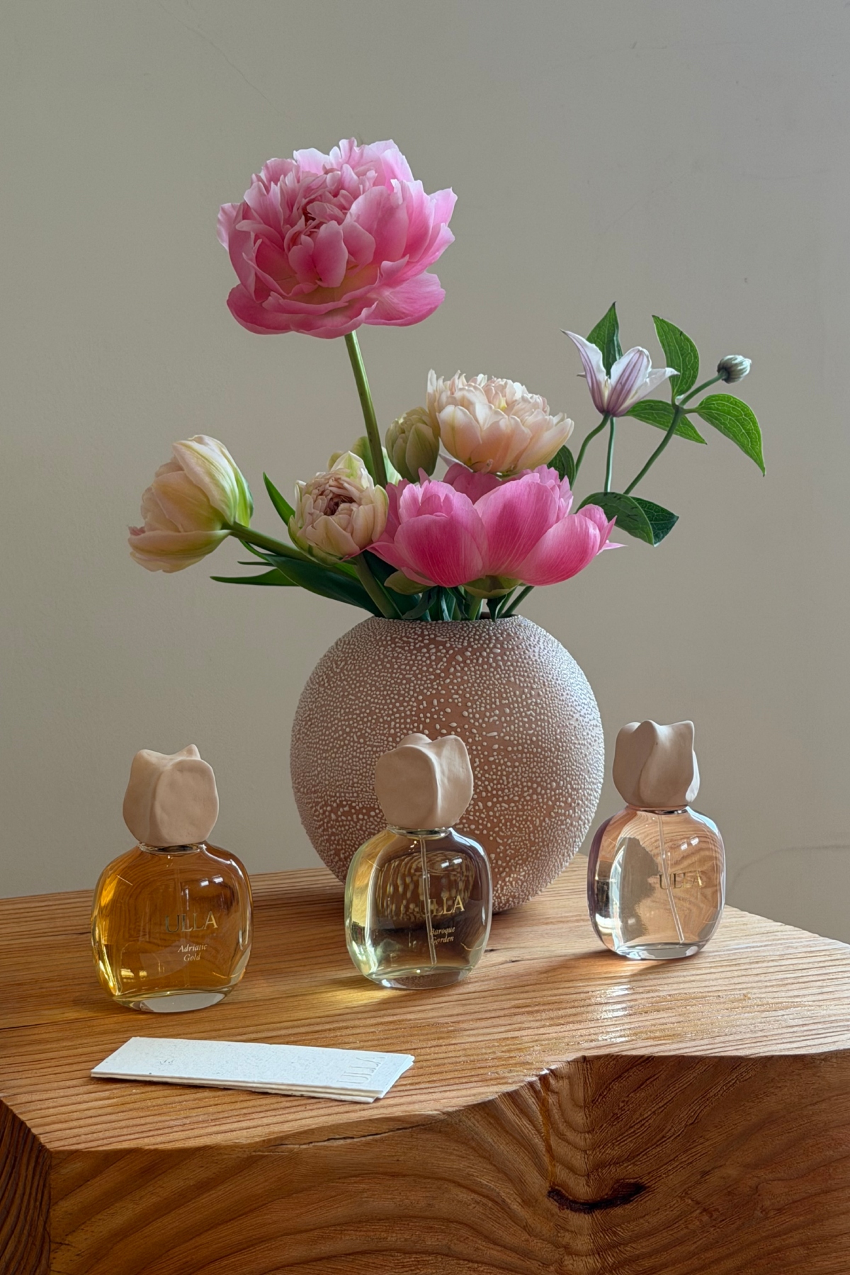 a round vase with pink and white flowers on a wood table with three glass bottles