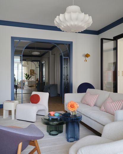 modern living room with parisian aesthetic with blue trimmed internal glass door, white sofa, white armchair, colored glass coffee tables, white rug, and sculptural pendant light