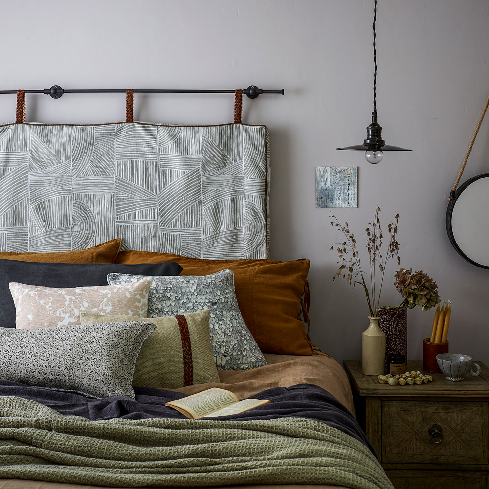 Grey bedroom with double bed, patterned cushions, wall hung headboard and wooden side table.