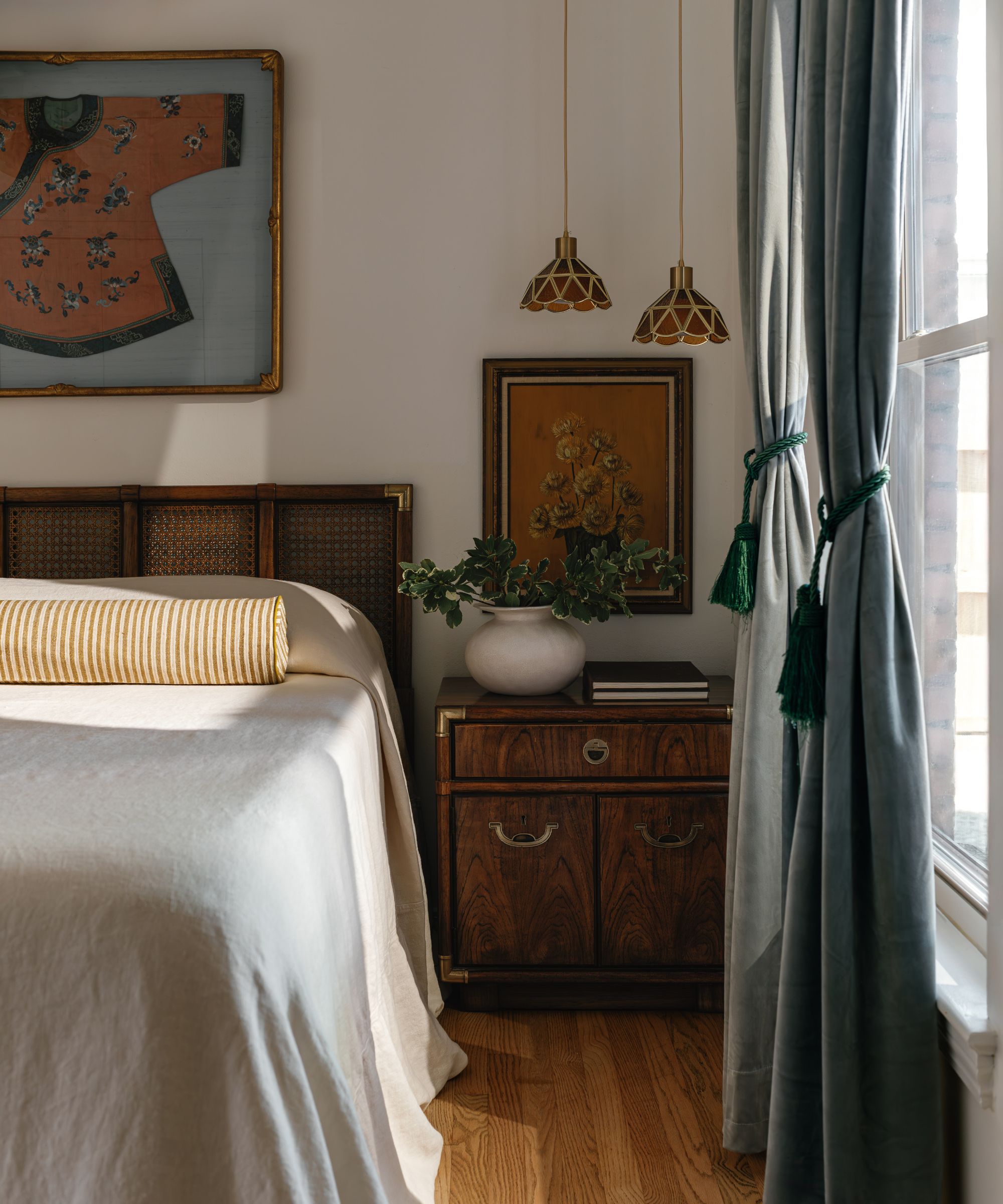 A bedroom with white walls, wooden flooring, and a wooden bed with an ochre bolster cushion, a dark wooden nightstand, and teal curtains.