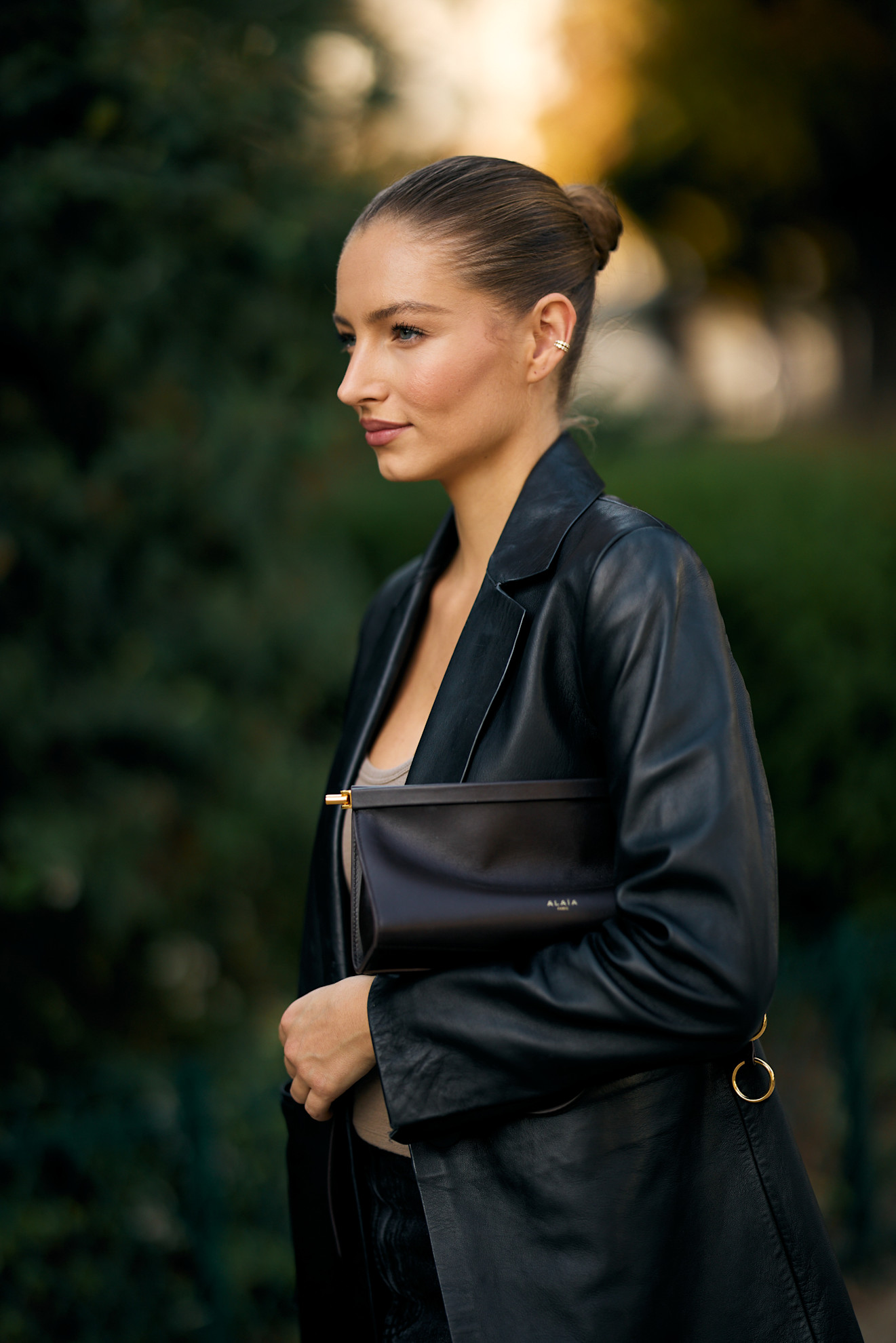 a white woman in a black leather jacket carrying a black leather clutch with a bun in her hair