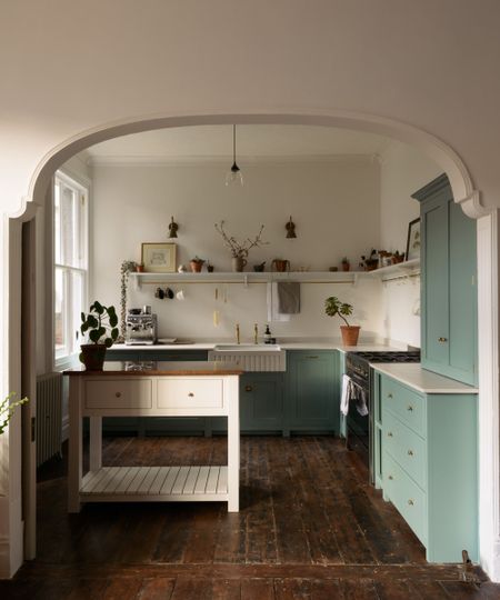 A small L-shaped kitchen with green cabinets, white countertops, an open shelf for decor, and a work table at the center acting as a kitchen island