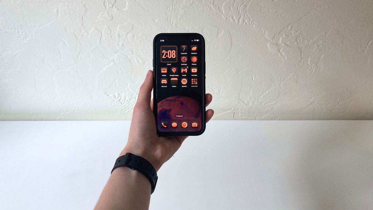 A person&#039;s hand holding an iPhone showing the home screen in iOS 18