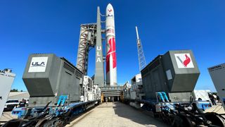 a red and white rocket rolls toward its launch pad with blue skies in the background.