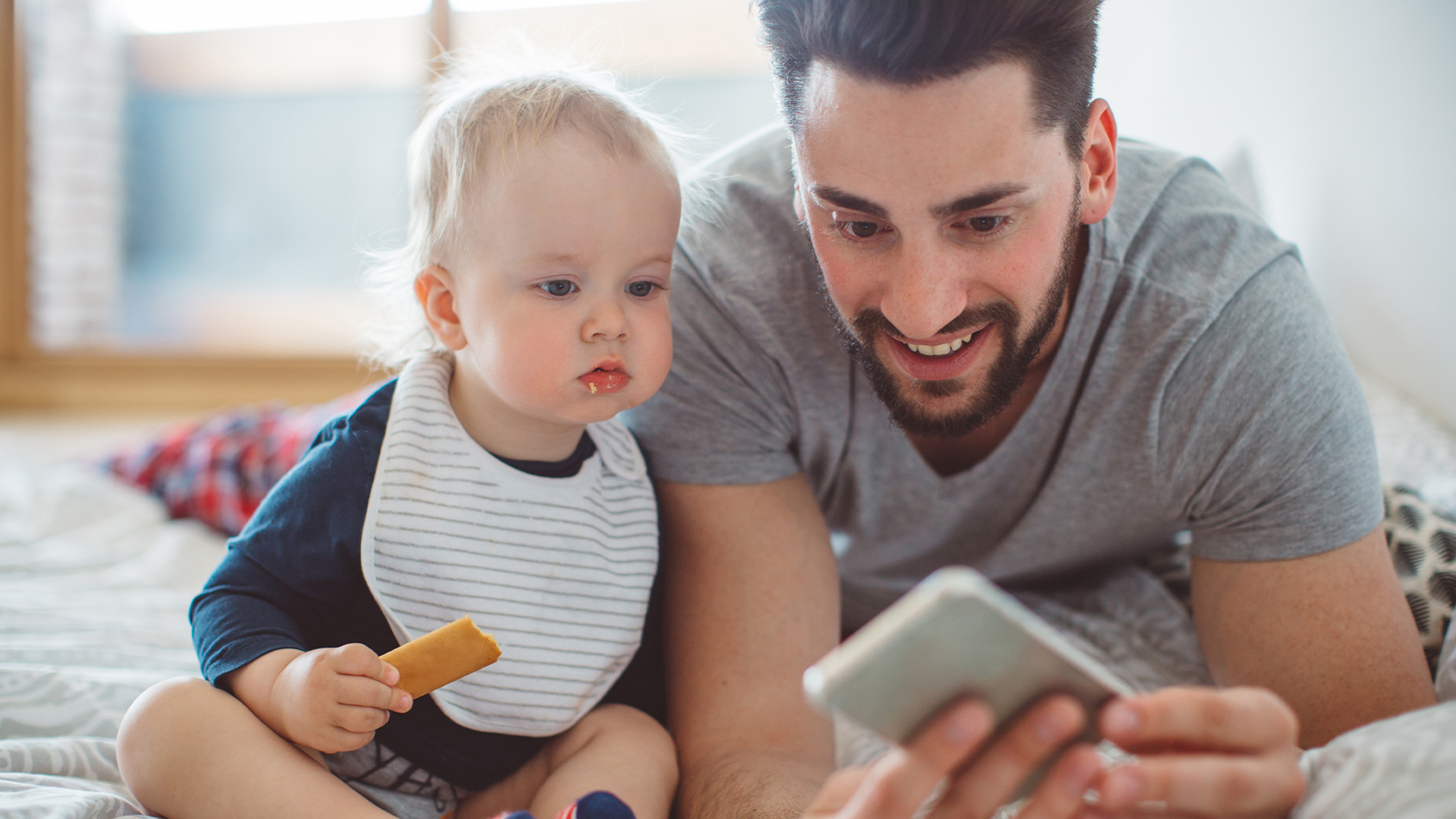 A man and a baby are next to each other on a bed, both looking at a smartphone