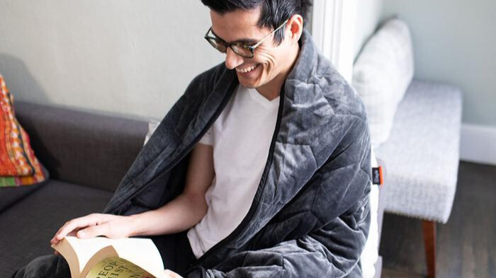 A man with a weighted blanket draped over him smiling while reading a book