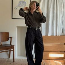 Woman takes a mirror selfie wearing pleated wide leg trousers, a brown button down and black belt.