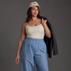Model wears white tank tucked in boxer style pants with baseball cap while draping a leather jacket over her shoulders