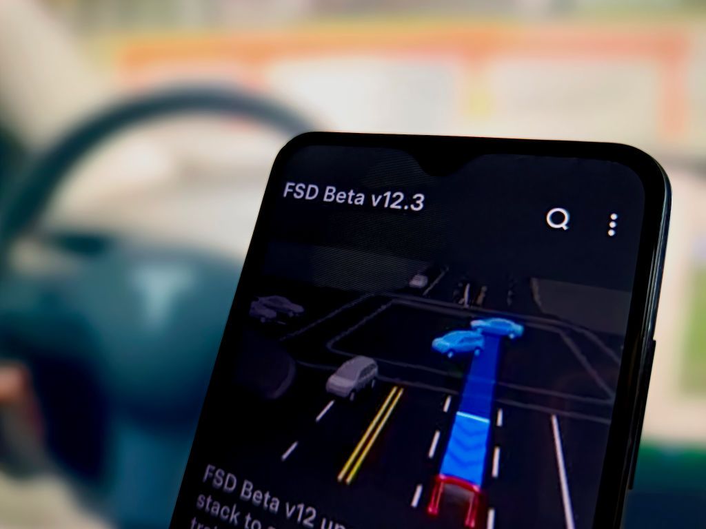 illustration of Tesla&#039;s FSD beta on smartphone with blurred steering wheel in background