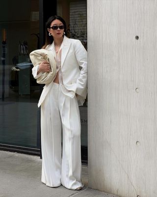 Woman wears all-white outfit with oversize blazer, white sheer shirt, and tailored pants, while holding white clutch and wearing black sunglasses, standing on street.