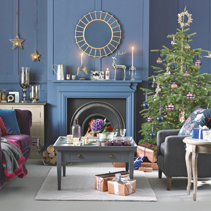 Blue living room with Christmas decoration, purple suede sofa, grey armchair, grey coffa table, decorated tree and fireplace.