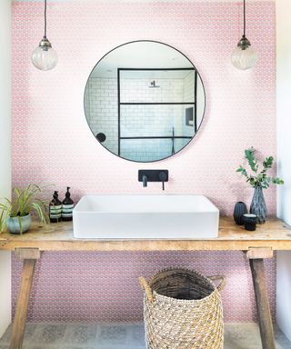 Pink penny tiles behind a white sink on a wooden table with a round mirror
