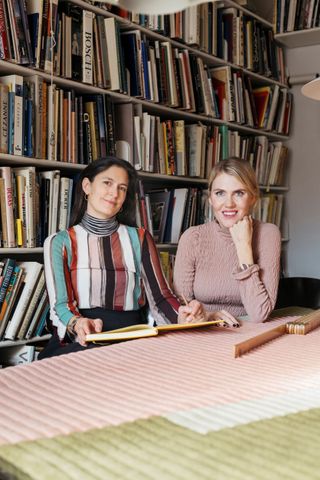 Image of Laura Juslin and Lilli Maunula sat together behind a table with pastel shade ribbed cloth, holding an open book, wooden measure, shelving with lots of books on, in the backdrop