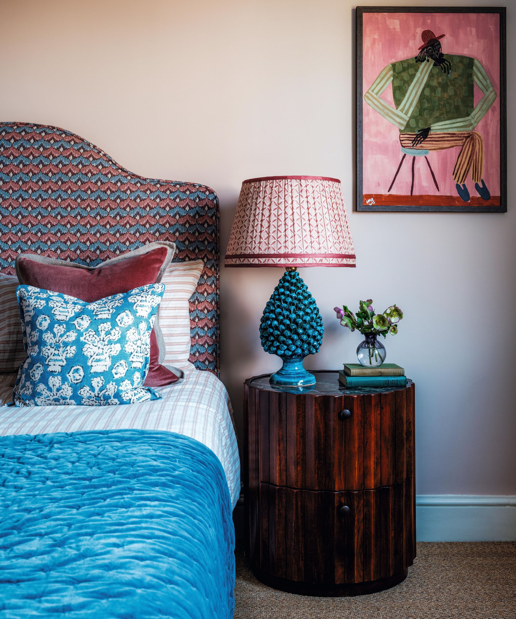 a primary bedroom painted muted pink with sisal carpet and a red and blue patterned headboard with matching mismatched linens and a dark wooden fluted nightstand