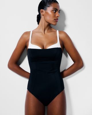 A Reformation model stands in front of a wall wearing a two tone one piece swimsuit
