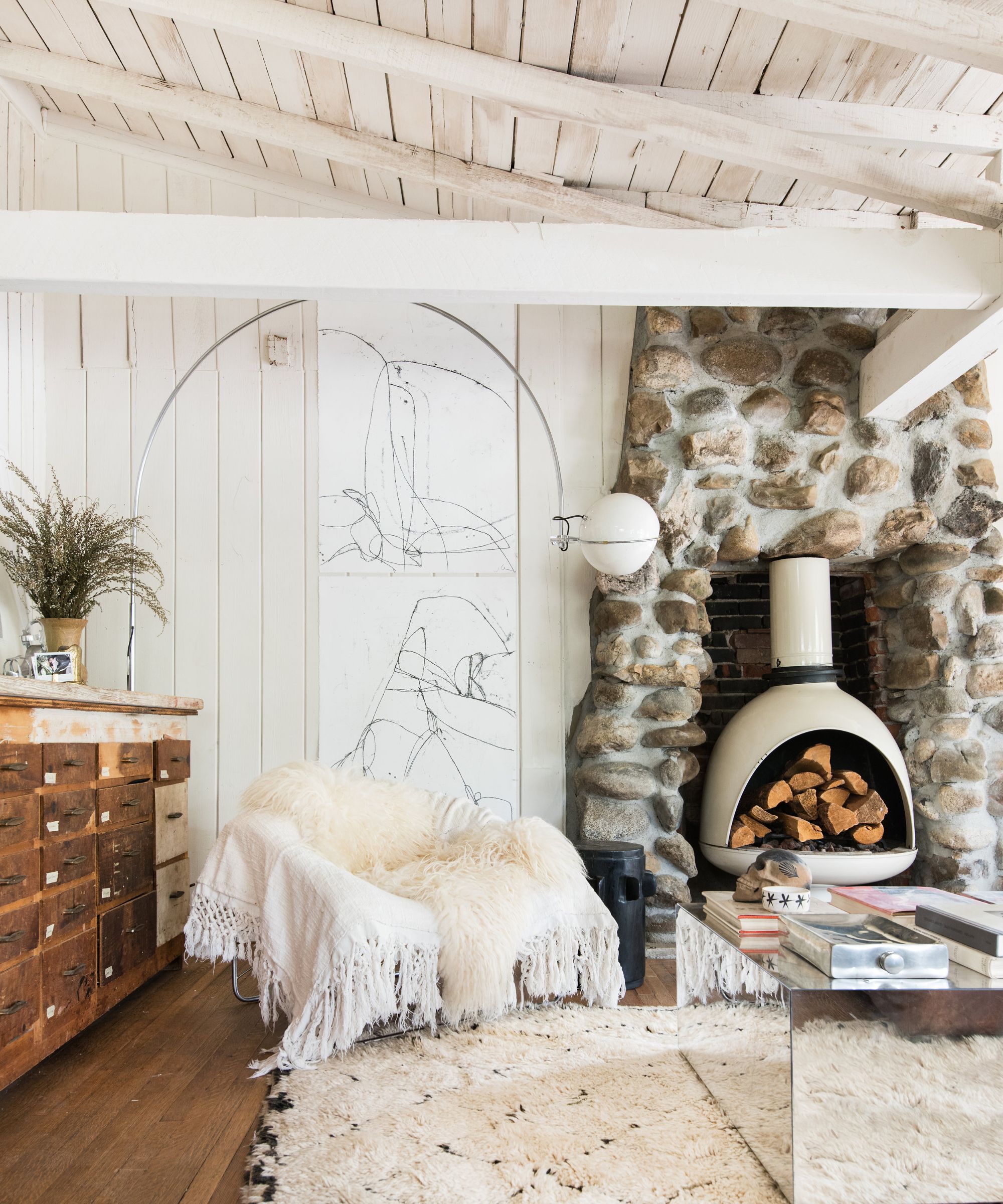 Living room with stone fireplace and log burner, white shiplap walls and beams, Berber rug, vintage wooden drawers, silver floor lamp, armchair with fluffy white throw and mirrored coffee table