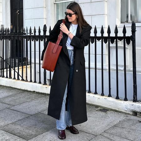 British influencer Anna Newton on a London sidewalk wearing a chic winter denim outfit with black sunglasses, a long black maxi coat, white t-shirt, straight-leg jeans, leather tote bag, and patent burgundy loafers