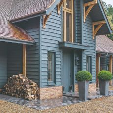 A blue house with a matching front door with two potted bushes framing the front porch and a pile of firewood to the side