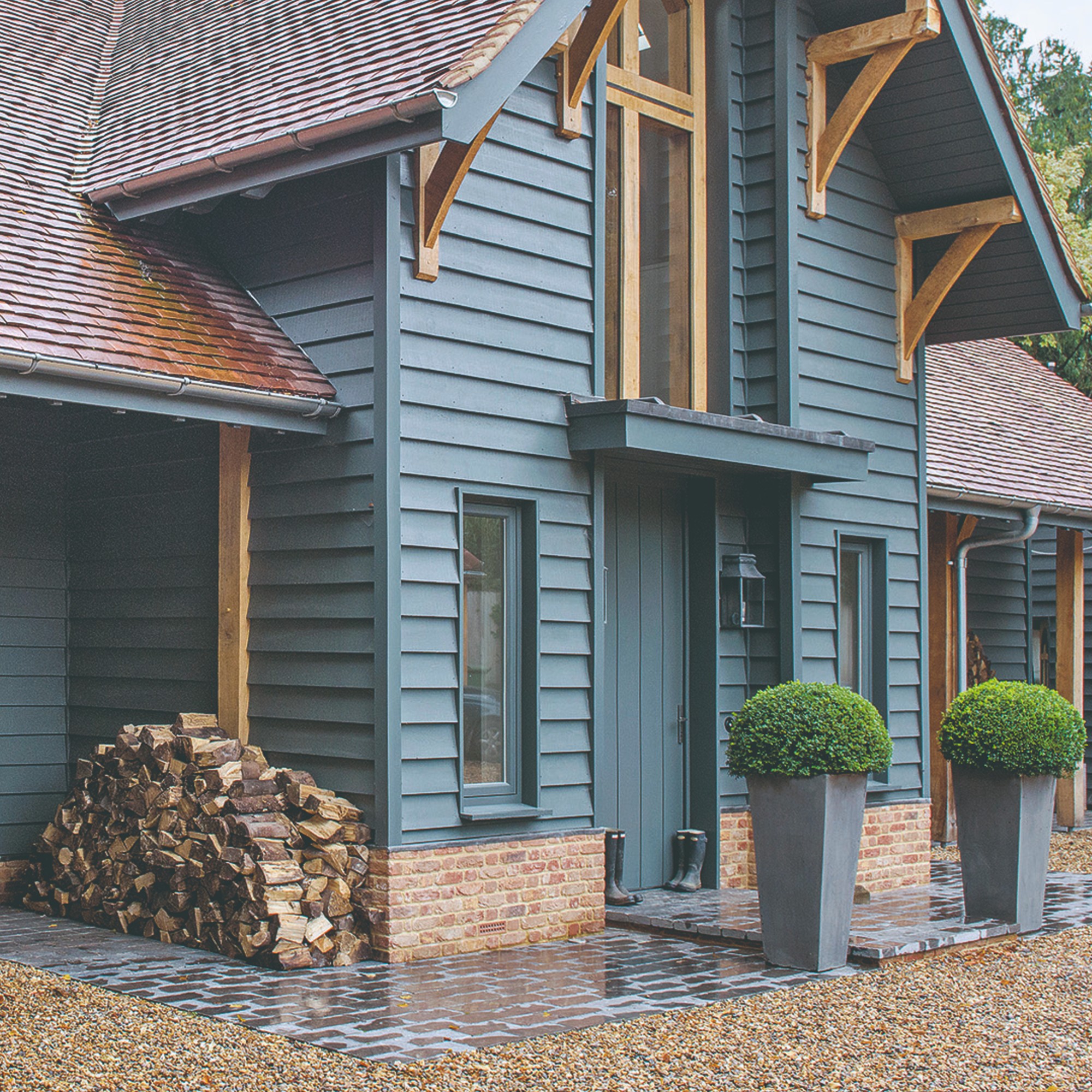 A blue house with a matching front door with two potted bushes framing the front porch and a pile of firewood to the side