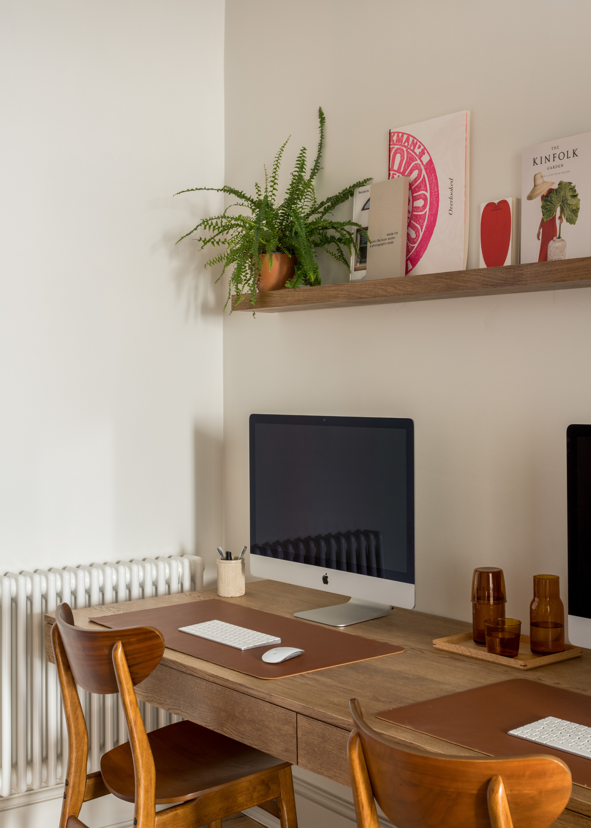 A white study nook with a mac on a wood desk with a pad for the mouse and keyboard, all below a floating shelf with books and a potted plant