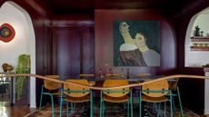 A maroon burgundy dining room with a large unframed painting, arched doorways, a wood table with wood and teal metal chairs, vases with flowers, and a trailing potted plant