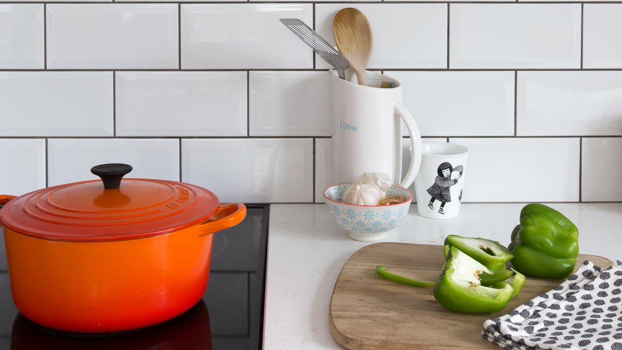 A white-tiled kitchen with a Le Creuset casserole dish on the hob and vegetables cut on a chopping board