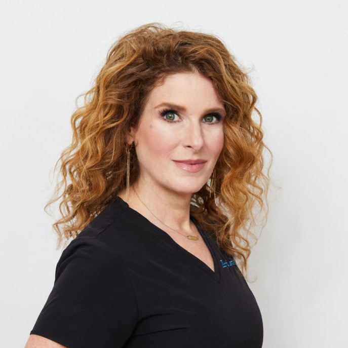 A woman, Dr. Amy B. Lewis, with curly red hair wearing medical scrubs. 
