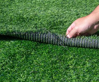 A mans hand lifting up the edge of fake grass to show the black mat underlayer