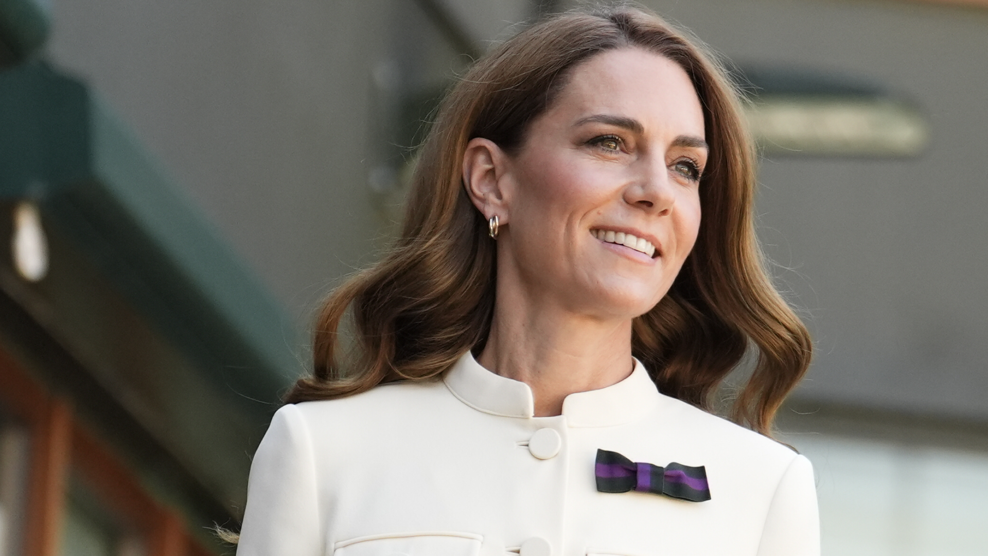 Kate Middleton wearing a white blazer with a purple bow pin and smiling