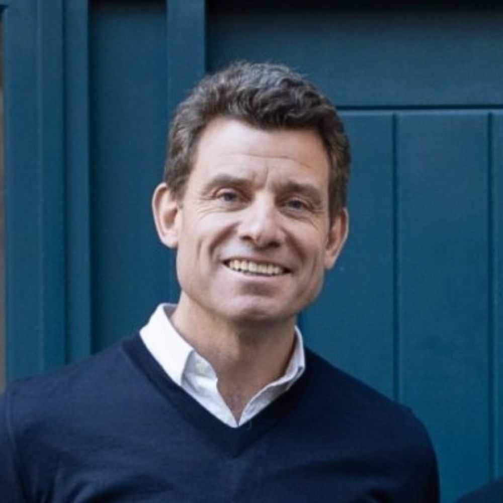 A smiling middle-aged man with short brown hair, wearing a navy V-neck jumper over a white shirt, photographed in front of a teal painted door.