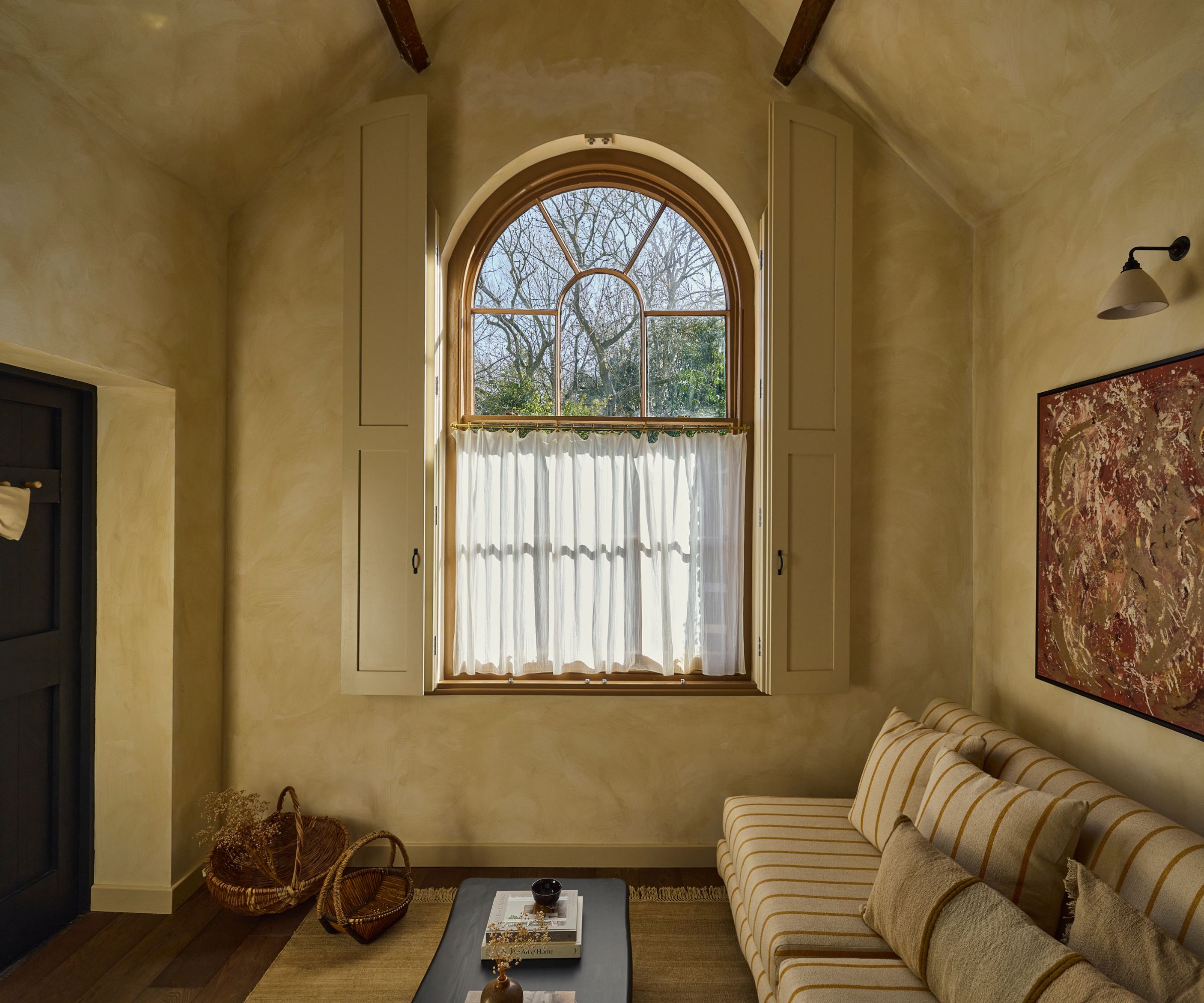 A tiny home living room with warm white limewash walls, a large arched window, a striped sofa, and earth-toned wall art