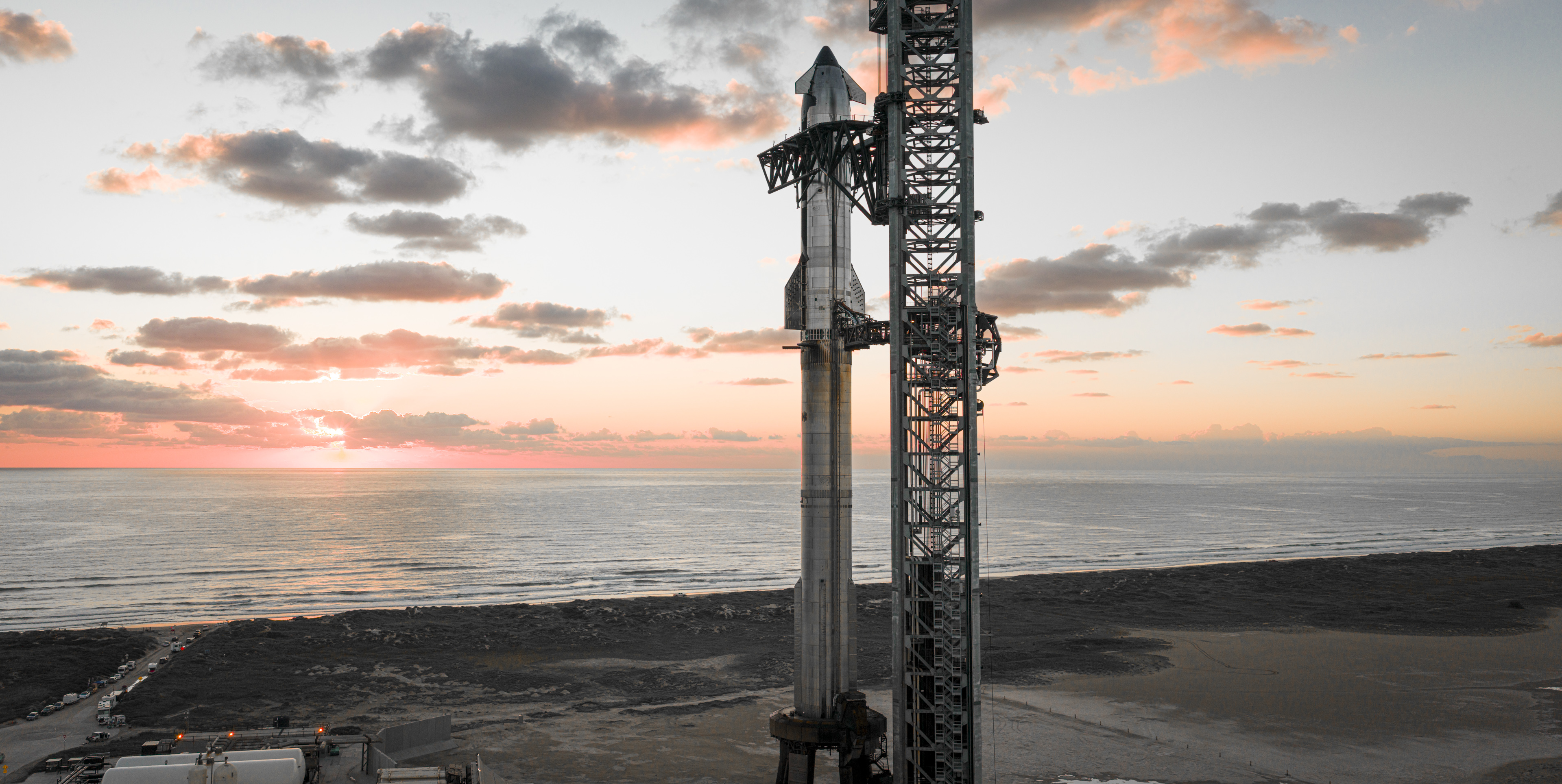 A giant silver SpaceX Starship rocket and its Super Heavy booster stand on the launch pad by the Gulf of Mexico with the sun on the horizon for Flight 11.