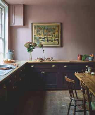 Black cabinets with pink walls, original wooden floorboards, artwork, table and chairs, wood countertop, vases, brass handles