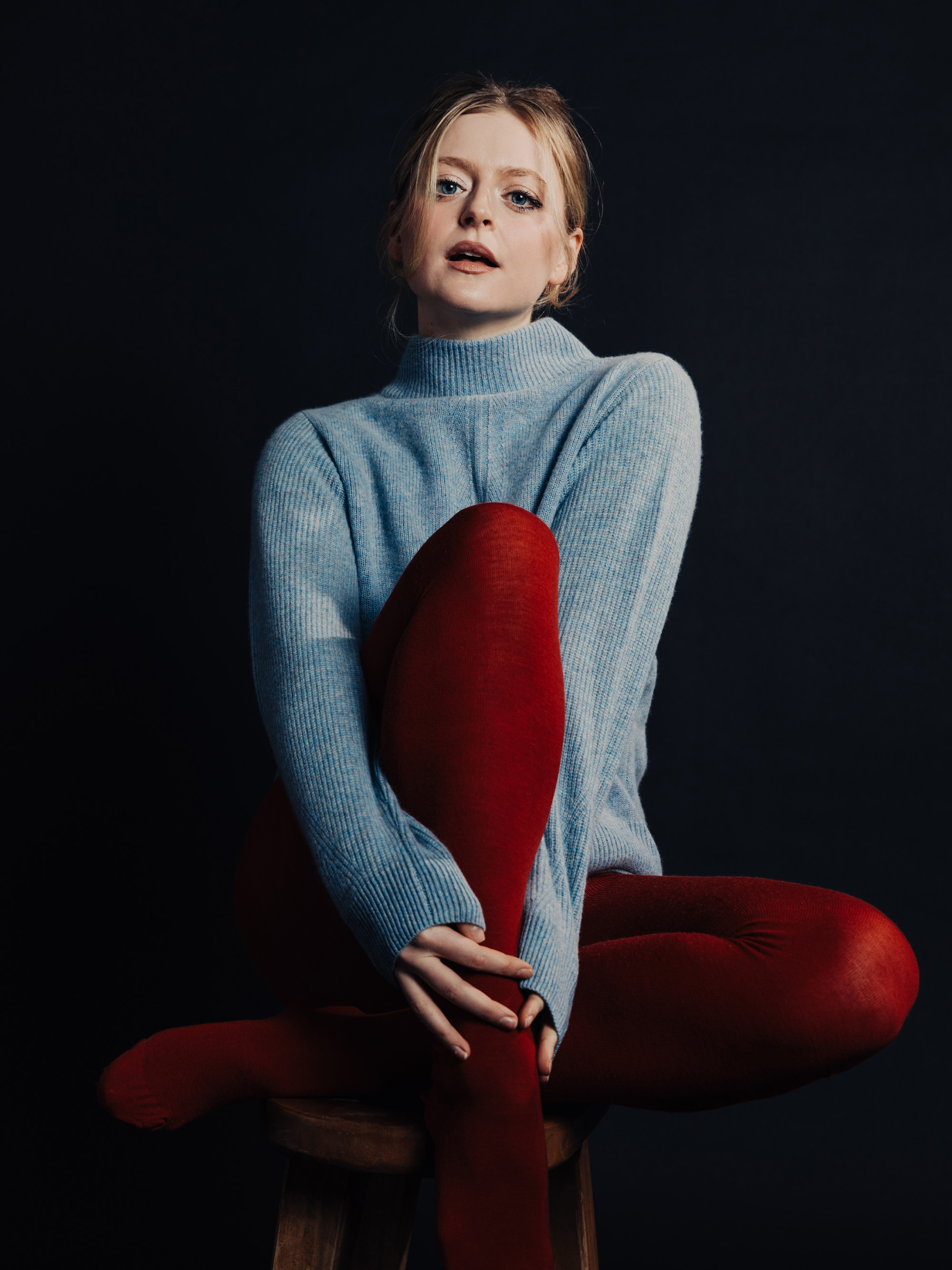 Actress Anna Baryshnikov photographed by Daniela Spector. She is wearing a baby blue mock neck sweater with red opaque tights. She is sitting on a stool against a black background.