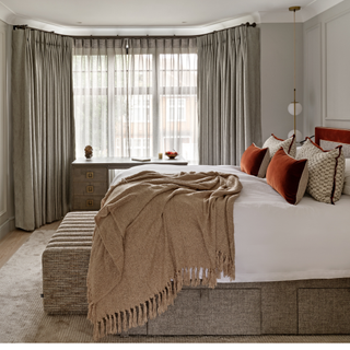 A bedroom with large bed covered in throws and cushions with a large bay window in the background dressed with curtains