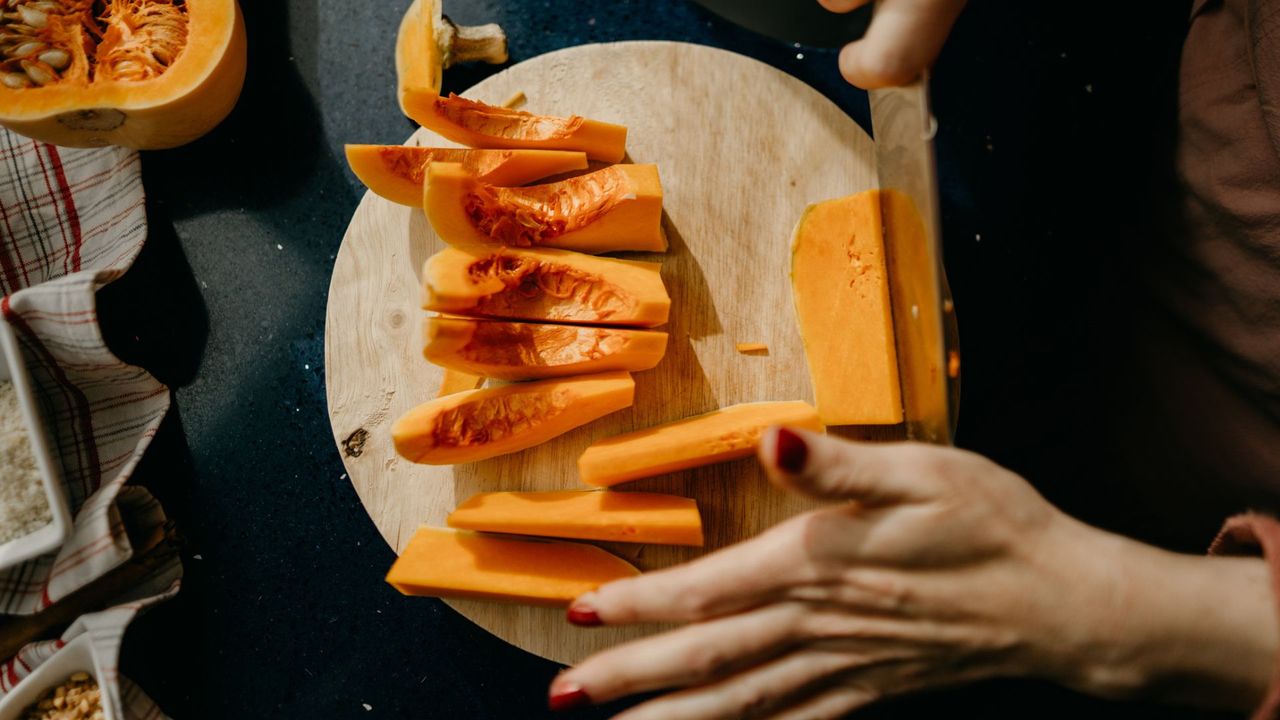 Woman&#039;s hands cutting up pumpkin on chopping board at kitchen counter, representing what to do with pumpkin seeds