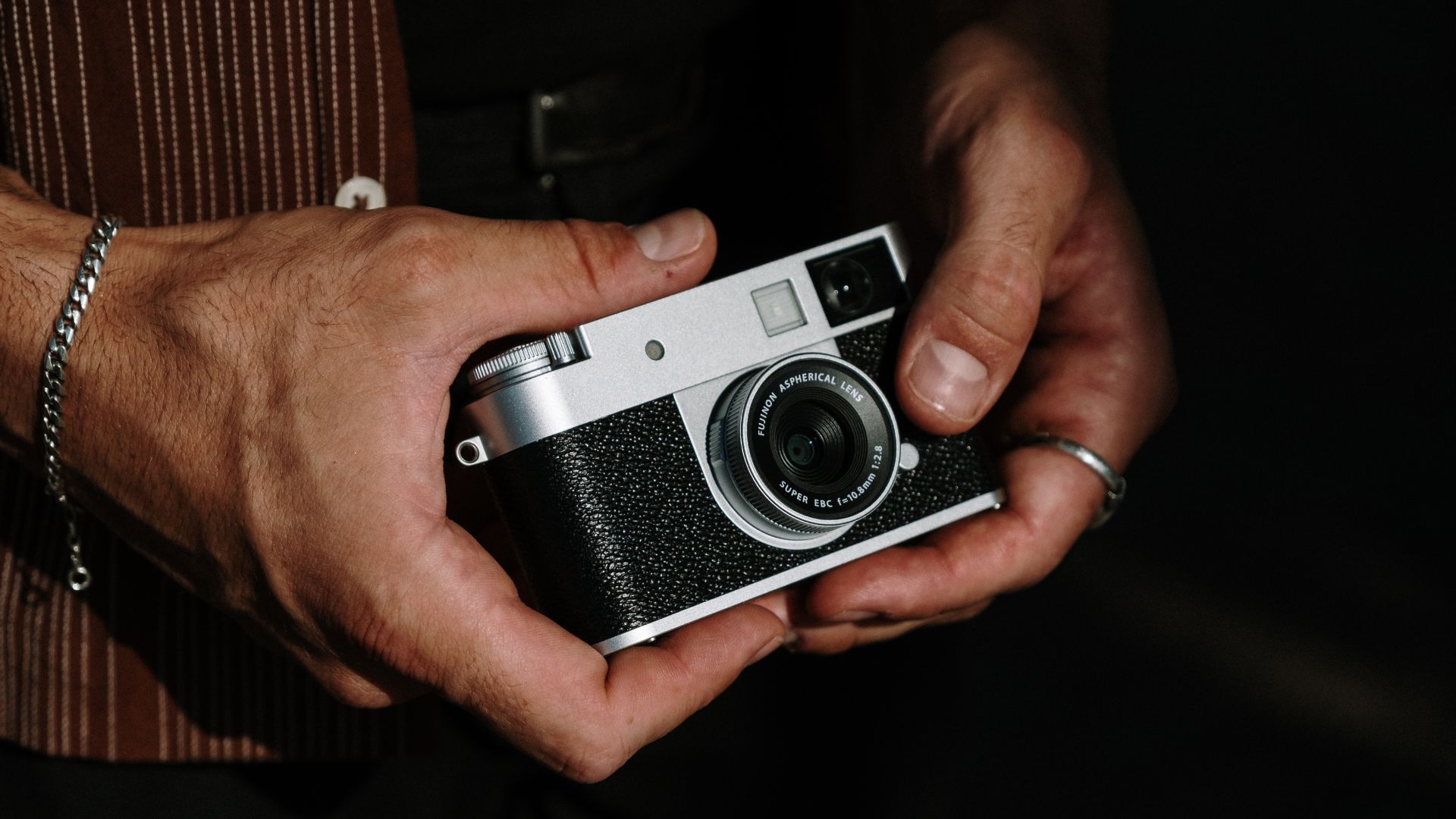 an image of a male model holding the Fujifilm half X camera between both hands. It is the silver colourway model and the man is wearing a brown striped shirt.