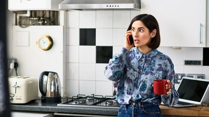 A woman on the phone in her kitchen looks anxious.