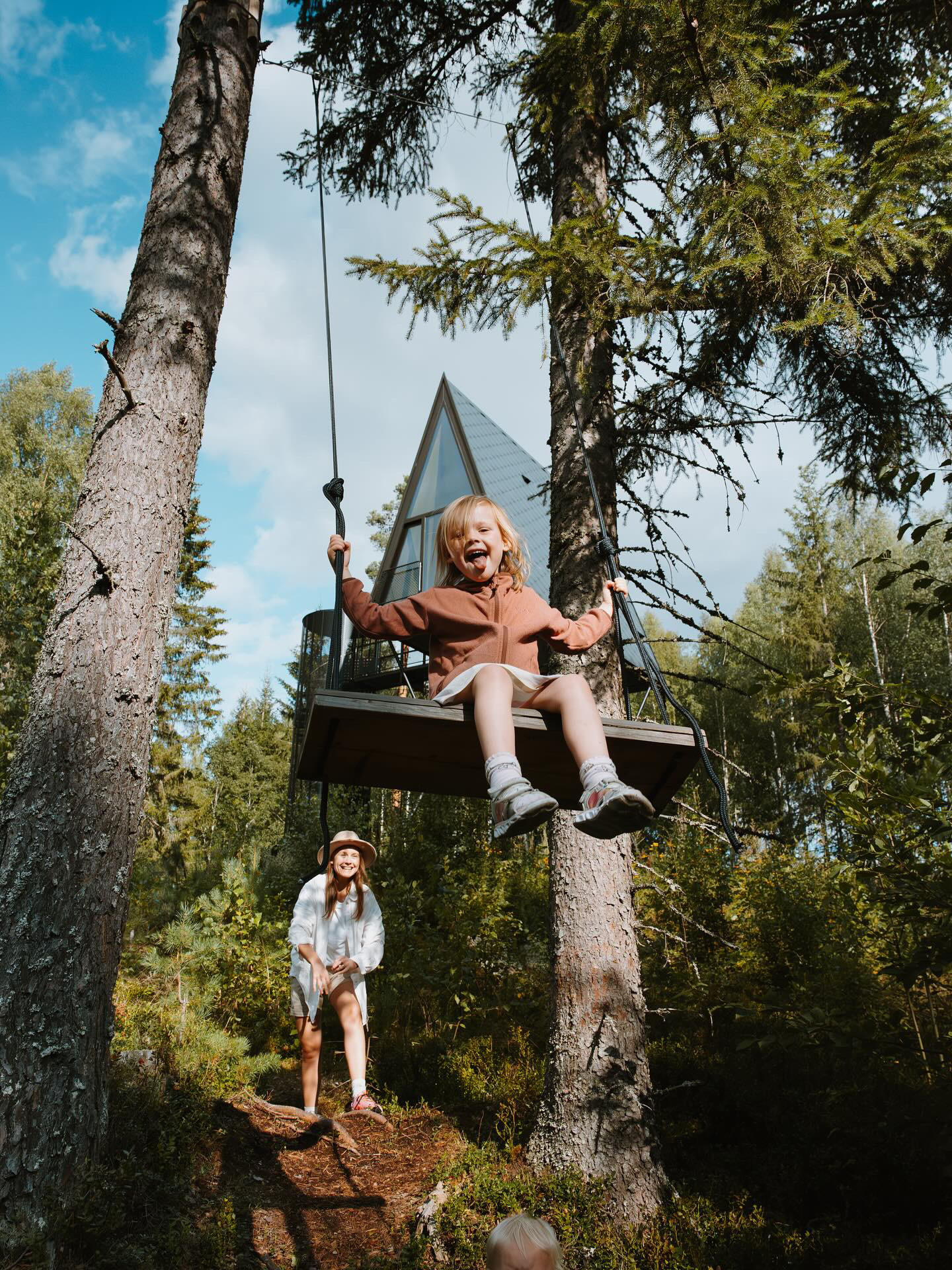 A mum and her young daughter play in a wood underneath a pointy design cabin, the little girl swinging on a swing, the mum pushing her on it.