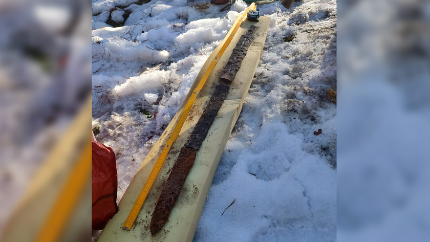 A corroded bronze sword broken into three parts is reassembled on a long light-colored wooden board. The board is on the snow-covered ground, and a yellow measuring tape is expanded beside the sword.